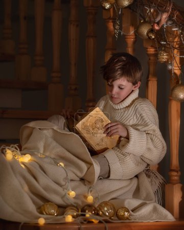 Téléchargez les photos : Garçon assis avec une fabuleuse boîte et des cadeaux près des escaliers à Noël. Nouvel An, bonheur et conte de fées - en image libre de droit