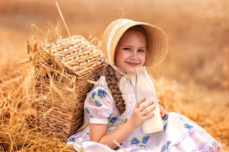 Foto de Un retrato de una chica feliz en un campo de trigo al atardecer. Un niño sostiene un frasco de vidrio con leche sobre el fondo de las orejas de centeno. Picnic en la naturaleza. - Imagen libre de derechos