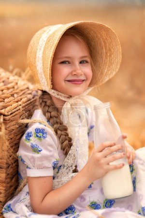 Foto de Un retrato de una chica feliz en un campo de trigo al atardecer. Un niño sostiene un frasco de vidrio con leche sobre el fondo de las orejas de centeno. Picnic en la naturaleza. - Imagen libre de derechos