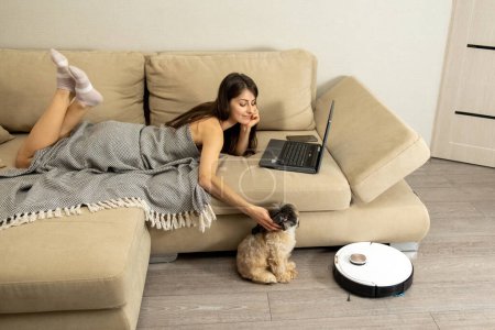 Photo for Covered with warm blanket young smiling woman lies on comfortable sofa and pets little cute dog. Shih Tzu enjoys sitting near robotic vacuum cleaner - Royalty Free Image