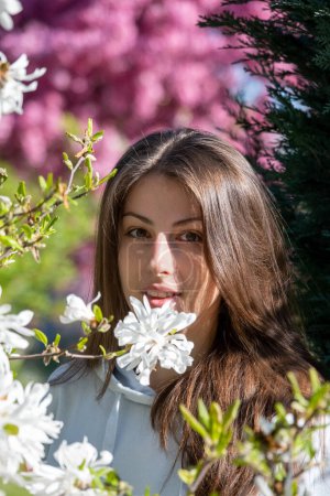 Téléchargez les photos : Jeune jolie femme pose près de l'arbre en fleurs dans le jardin botanique. Dame aux cheveux bruns souriant regarde droit jouissant parfum de printemps à l'extérieur gros plan vertical - en image libre de droit