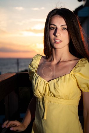 Téléchargez les photos : Portrait de jeune femme brune posant contre le coucher du soleil d'été. Brunette dame en robe jaune jouit brise de mer debout sur le balcon de l'hôtel de luxe vertical - en image libre de droit