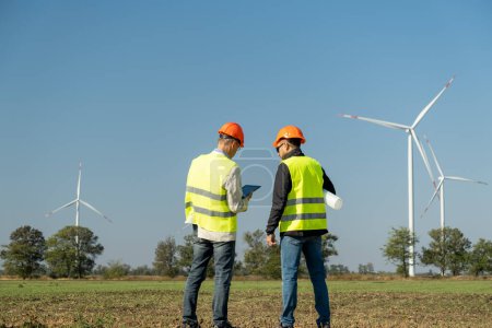 Photo for Windmills produce alternative energy in countryside. Technicians of maintenance in helmets talk about project against wind generators back view - Royalty Free Image
