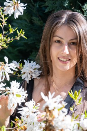 Foto de Mujer sonriente joven se para cerca de un árbol floreciente de primavera en el campo. Señora de pelo oscuro disfruta de la fragancia de las flores blancas de primavera en el primer día soleado vertical - Imagen libre de derechos