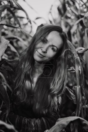 Foto de La mujer se para y disfruta explorando las plantaciones de maíz en el campo. La hembra de pelo largo mira y toca hojas de callos - Imagen libre de derechos