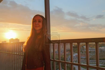 Téléchargez les photos : Femme se penche sur la clôture du balcon et admire les moments au soleil arrière. Coucher de soleil traverse les nuages au-dessus des immeubles de grande hauteur et illumine la ville - en image libre de droit