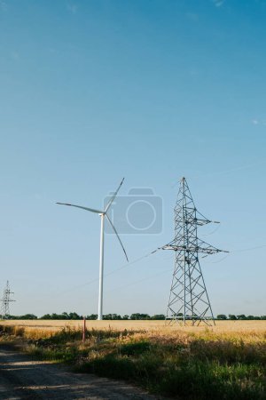 Photo for Windmill producing renewable energy. Ecofriendly wind turbine generates clean energy near power transmission line on wheat field illuminated by sun - Royalty Free Image