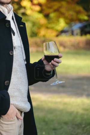 Téléchargez les photos : Homme méconnaissable en vêtements chauds détend boire du vin rouge en vacances. élégant gars se tient sur gazon pelouse tenant la main dans la poche sur fond flou - en image libre de droit