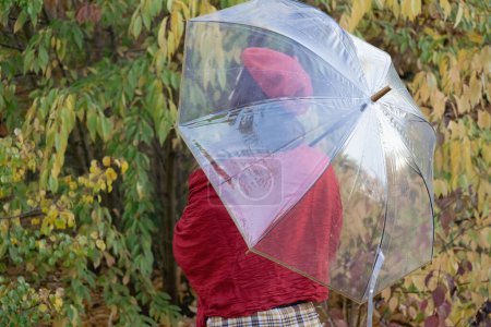 Téléchargez les photos : KYIV, UKRAINE - 27 SEPTEMBRE 2021 : Une femme méconnaissable regarde de côté tenant un parapluie transparent contre la forêt d'automne. Dame aux cheveux foncés en écharpe rouge jouit d'une vue arrière campagne pluvieuse - en image libre de droit