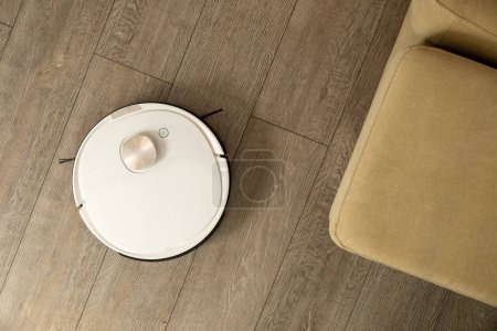 Foto de La aspiradora robótica blanca funciona en la sala de estar ligera. Dispositivo moderno limpia piso laminado cerca de sofá beige en primer plano apartamento vista superior - Imagen libre de derechos