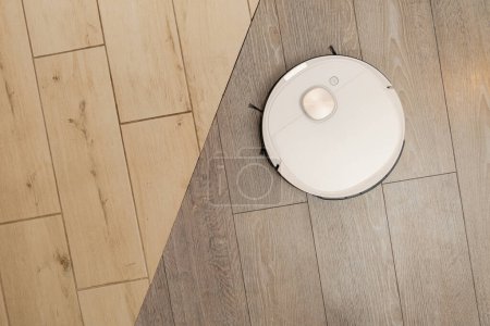 Téléchargez les photos : Aspirateur robotique autonome blanc nettoie les sols carrelés et stratifiés à motifs de bois. Dispositif pour la collecte commode de poussière et de saleté dans l'appartement vue rapprochée supérieure - en image libre de droit