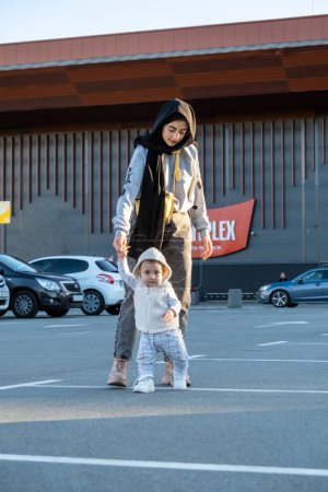 Foto de Madre musulmana camina cerca del mercado y se encarga de niño emocionado. Niño con expresión divertida y alegre disfruta caminar con los padres en el estacionamiento - Imagen libre de derechos