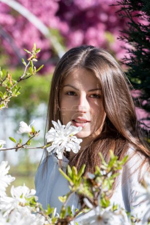 Photo for Portrait of brunette lady standing near young branches of white blossoms in spring park. Young woman unites with nature looking straight vertical - Royalty Free Image