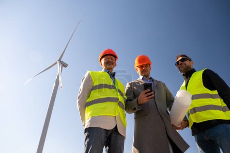 Photo for Wind turbine rotates behind gathered specialists on sunny day. Chief architect and engineers check operation of built wind farm low angle shot - Royalty Free Image