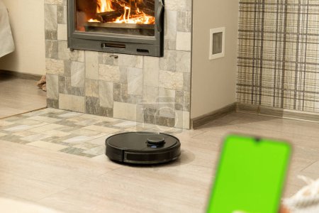 Photo for Smartphone screen with chromakey against cozy light room. Modern black robotic vacuum cleaner cleans tiled floor near fireplace in apartment closeup - Royalty Free Image