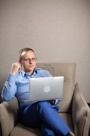 Téléchargez les photos : Psychologue homme dans les lunettes regarde attentivement à l'écran d'ordinateur portable. Homme tient le dispositif sur le genou et s'assoit dans le fauteuil dans le bureau léger - en image libre de droit