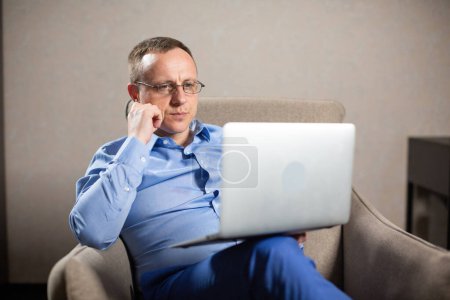 Téléchargez les photos : Psychologue masculin joue sac à main et regarde ordinateur portable. Homme tient le dispositif sur le genou et s'assoit dans le fauteuil dans un local spacieux - en image libre de droit