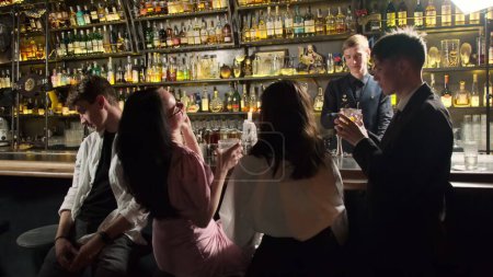 Téléchargez les photos : KYIV, UKRAINE - 27 SEPTEMBRE 2021 : Un homme fatigué est assis au comptoir du bar, loin de la compagnie bruyante. Ami joyeux faire des toasts avec barman préparer des cocktails en arrière-plan - en image libre de droit