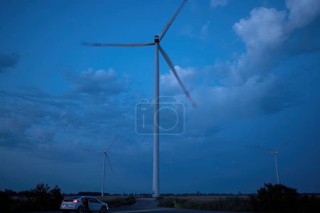 Foto de Molino de viento genera energía limpia contra el cielo azul al atardecer. Generador de viento ecológico produce energía renovable y coche con los turistas explorando proceso - Imagen libre de derechos
