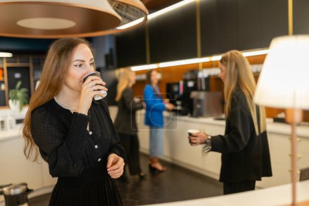 Photo for Young women share small talk and drink hot beverages in office canteen. Female employees communicate and sip warm drinks during work break - Royalty Free Image