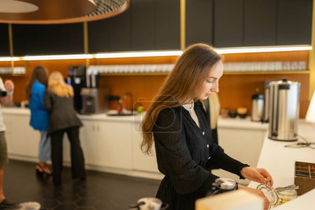 Photo for Young businesswoman makes hot beverage to recharge during work break in staff canteen. Female employees prepare hot drinks in workplace kitchen - Royalty Free Image