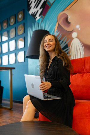 Téléchargez les photos : KYIV, UKRAINE - 27 SEPTEMBRE 2021 : Femme souriante assise sur un canapé rouge et tenant un ordinateur portable. Femme agréable en tenue noire se trouve par dispositif d'argent moderne et regarde dans la caméra - en image libre de droit
