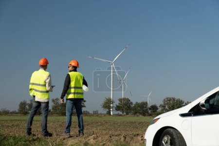 Téléchargez les photos : Les éoliennes produisent de l'énergie pour les voitures électriques dans les campagnes. Les techniciens d'éoliennes en casques orange se dressent contre de puissants moulins à vent - en image libre de droit