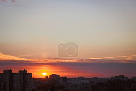 Photo for Sun sets down and illuminates evening city silhouettes with last rays breaking through clouds in sky. Multi-storey buildings rise above dark trees - Royalty Free Image