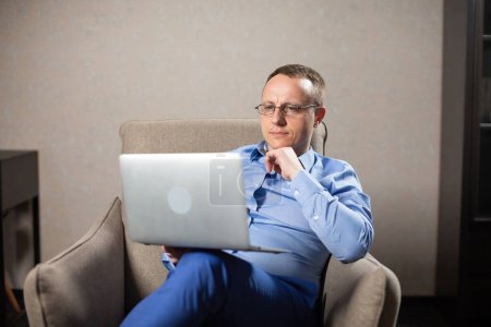 Photo for Thoughtful psychologist thinks about solving problems of patient. Man sitting in armchair with white laptop on lap in room of apartment - Royalty Free Image