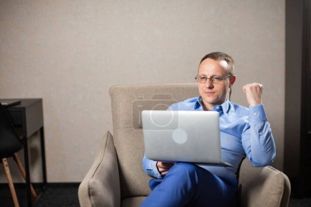 Photo for Male psychologist of company sitting in grey armchair and looking at screen of laptop. Busy man conduct online appointment in cozy premise - Royalty Free Image