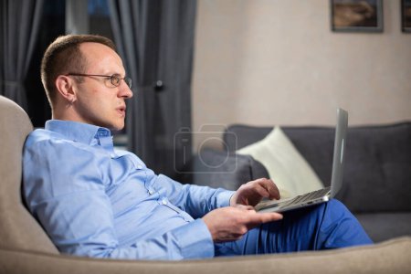 Photo for Thoughtful psychologist thinks about solving working tasks. Male doctor sitting on armchair with white laptop on lap in light premise - Royalty Free Image