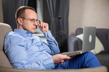 Photo for Serious psychologist with glasses sitting in grey armchair and working on laptop. Male doctor reads patient history in light premise - Royalty Free Image