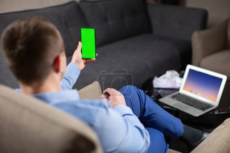Téléchargez les photos : Psychologue masculin assis sur le lieu de travail et regardant l'écran de téléphone portable avec chromakey vert. Ordinateur portable avec image sur le bureau - en image libre de droit