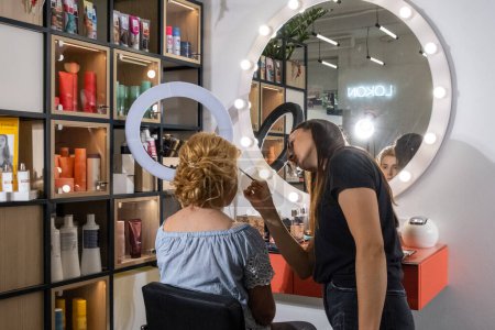 Téléchargez les photos : KYIV, UKRAINE - 27 SEPTEMBRE 2021 : Styliste maquille un client près d'un grand miroir rond dans un salon de beauté. Maquilleuse penche vers la cliente avec le pinceau à la main - en image libre de droit