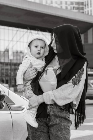 Téléchargez les photos : KYIV, UKRAINE - 27 SEPTEMBRE 2021 : Femme regarde adorable tout-petit fils dans les bras exprimant l'amour à l'enfant. Dame portant hijab noir aime marcher en plein air avec l'enfant et explorer le monde - en image libre de droit