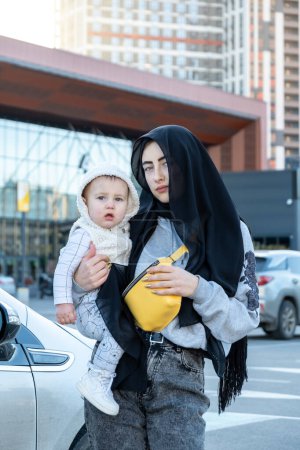 Téléchargez les photos : KYIV, UKRAINE - 27 SEPTEMBRE 2021 : Femme portant le hijab noir tient l'enfant dans les bras contre le stationnement occupé et aime prendre soin de tout-petit. Adorable fils regarde autour avec amusement - en image libre de droit