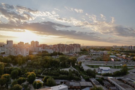 Foto de El atardecer ilumina la ajetreada ciudad con zona residencial rodeada de parque con árboles verdes y carretera. La luz del sol atraviesa las nubes de cúmulos - Imagen libre de derechos