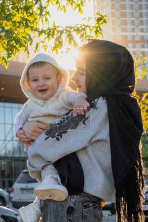 Foto de KYIV, UCRANIA - 27 DE SEPTIEMBRE DE 2021: Emocionado niño en brazos de madre quiere jugar en la calle a la luz del sol. Adorable niño quiere caminar en el estacionamiento con la mujer en hijab - Imagen libre de derechos