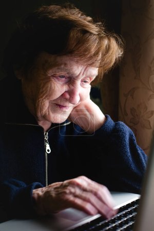 Téléchargez les photos : Une femme âgée est assise sur un ordinateur portable, son visage illuminé par la douce lueur de l'écran. Avec une concentration concentrée, elle tape loin, ses mains ridées se déplaçant sur le clavier. - en image libre de droit