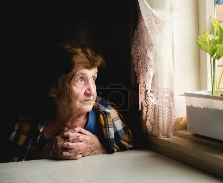 Une femme âgée assise près de la fenêtre, regardant dehors avec une expression réfléchie, perdue dans la contemplation.