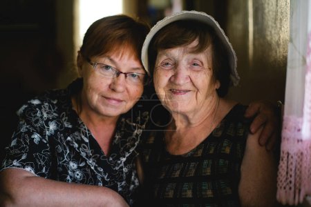Eine ältere Frau und ihre erwachsene Tochter teilen einen zärtlichen Moment im Porträt, der den Lauf der Zeit und die bleibende Verbundenheit zwischen den Generationen widerspiegelt..