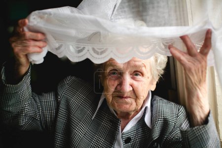 Une dame âgée regarde à travers un rideau de dentelle, ses yeux remplis d'histoires et de souvenirs.