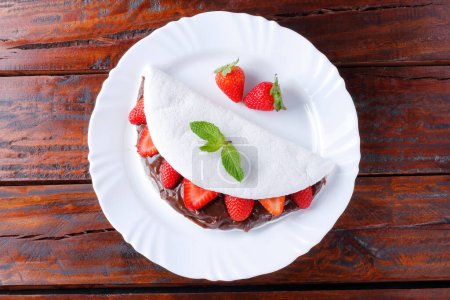 hausgemachte Tapioka oder Beiju gefüllt mit Erdbeere und Schokolade auf weißem Teller über rustikalem Holztisch. Ansicht von oben