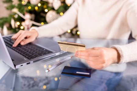 Photo for Cropped shot of female hands with golden credit card and laptop. Christmas presents, decorated tree on background. Woman shopping for holiday season presents online. Wooden table, copy space, close up - Royalty Free Image