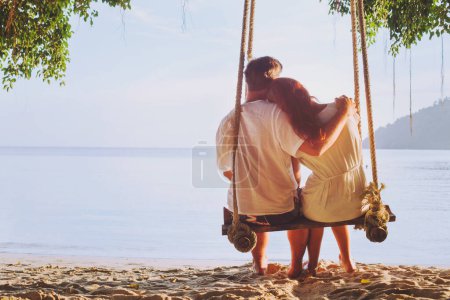Foto de Vacaciones románticas para dos, pareja cariñosa sentados juntos en la playa en el columpio, silueta de hombre abrazando a mujer - Imagen libre de derechos