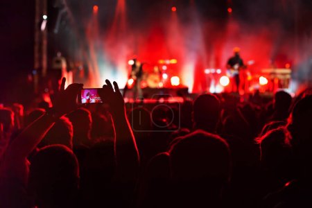 hands filming concert with smartphone, crowd of spectators watching musicians playing music and singing on stage