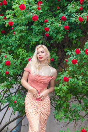 Téléchargez les photos : Belle femme aux longs cheveux blonds et aux lèvres rouges portant des vêtements roses pose près des roses en fleurs dans le jardin. Portez un haut et une jupe rose clair - en image libre de droit