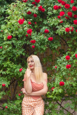 Téléchargez les photos : Belle femme aux longs cheveux blonds et aux lèvres rouges portant des vêtements roses pose près des roses en fleurs dans le jardin. Portez un haut et une jupe rose clair - en image libre de droit