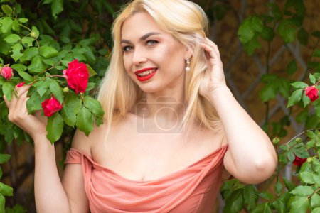 Photo for Beautiful woman with long blonde hair and red lips wearing pink clothes poses near blooming roses in garden. Wear light pink top and skirt - Royalty Free Image