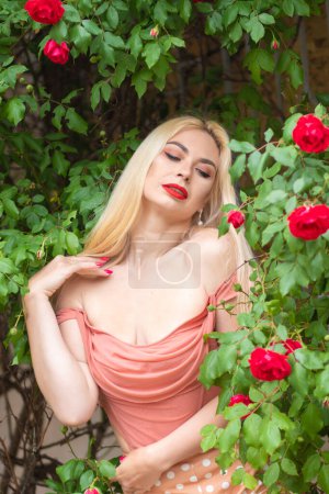 Photo for Beautiful woman with long blonde hair and red lips wearing pink clothes poses near blooming roses in garden. Wear light pink top and skirt - Royalty Free Image